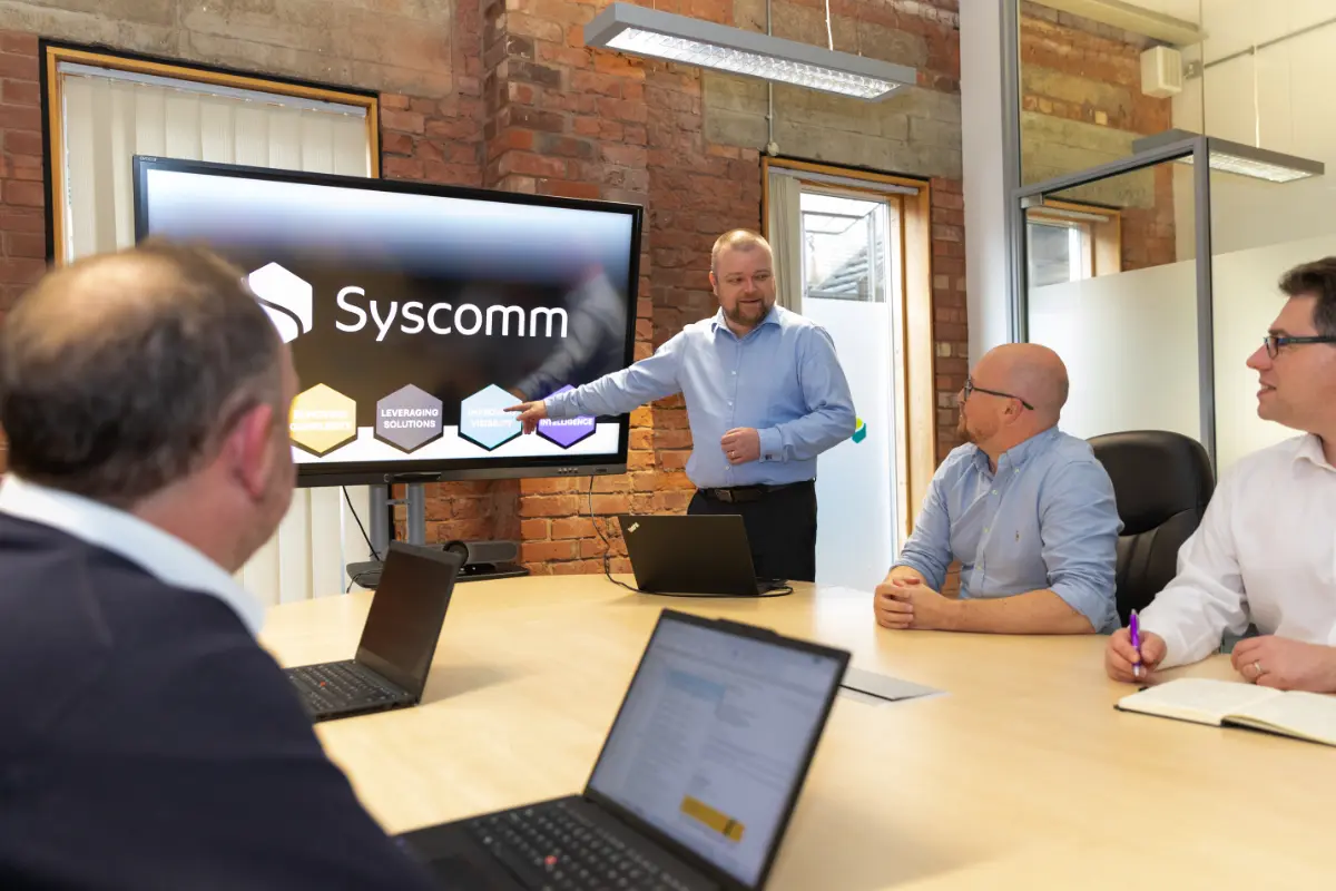 Business meeting in a modern office with a presenter pointing to a screen displaying the Syscomm logo
