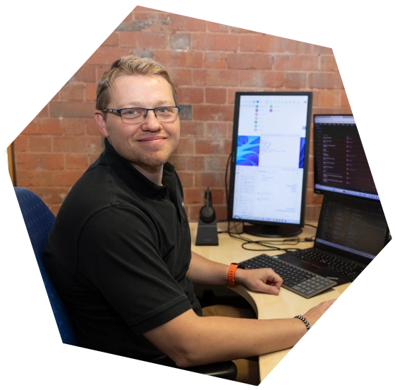 Man wearing glasses and a black polo shirt smiling at the camera while working at a desk with two monitors