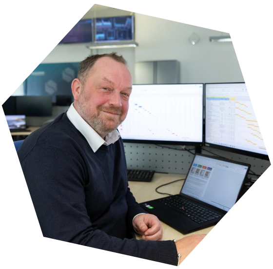 Smiling man in a navy sweater sitting at a desk with multiple monitors displaying charts