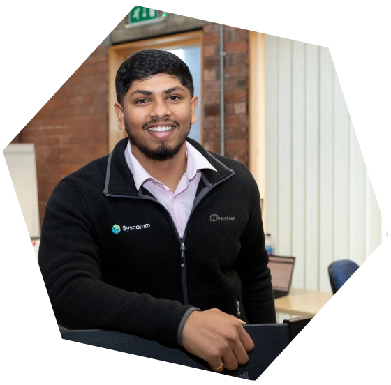 Smiling man in branded Synccomm and DrayTek jacket standing in a modern office environment