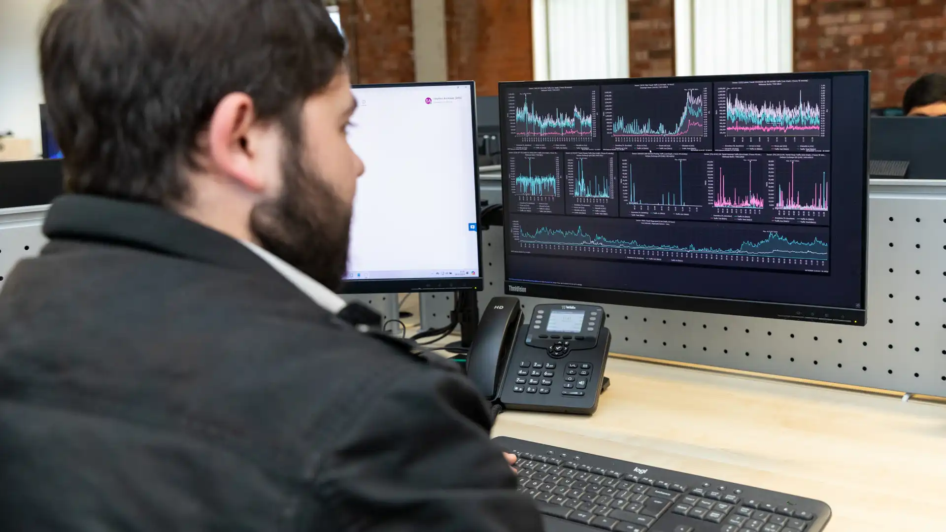 man in a black jacket working at a desk with two monitors displaying network performance analytics